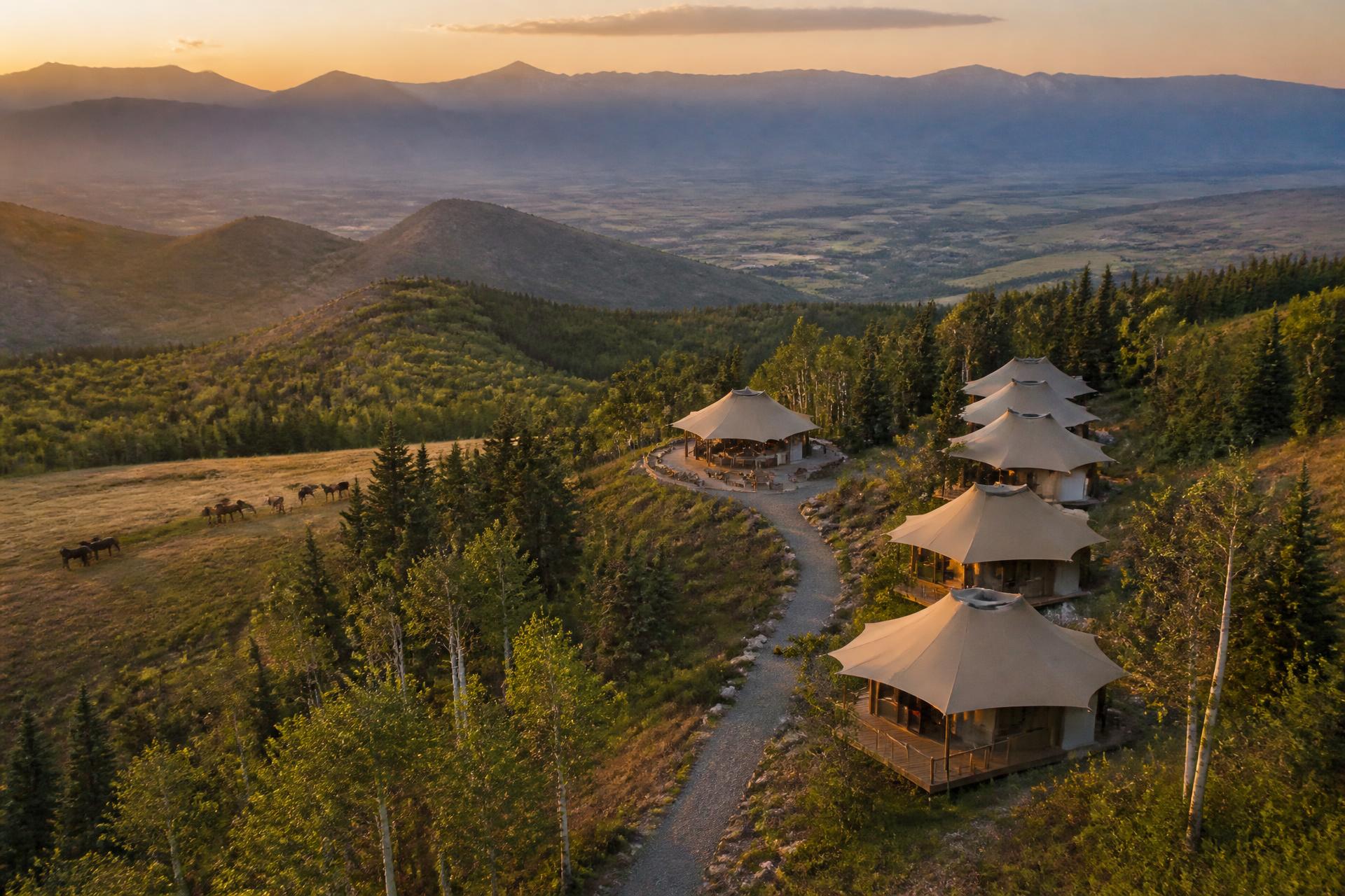 Suites strung along the ridgeline in the aspen and pine, gathering toward a central pavilion — horses in the meadow below, valley opening beyond.