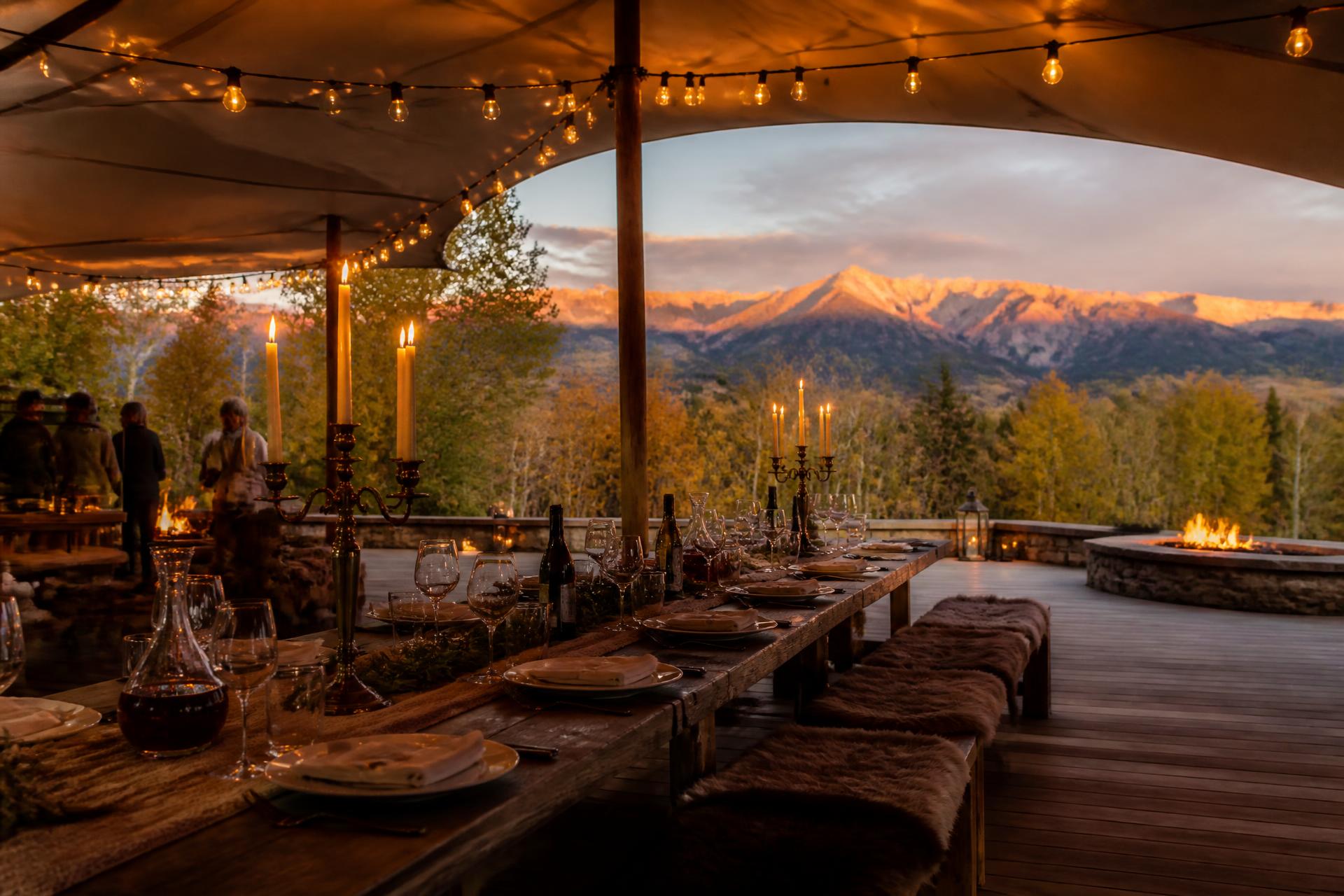 The central pavilion at dusk — long timber table under tensile canvas, candelabra and lantern light held against the last fire on the peaks.